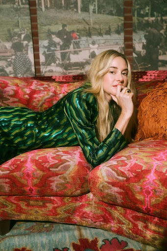 Adult female with blonde hair lies on a patterned sofa indoors, wearing a shiny green patterned dress, hand on chin, looking at the camera.