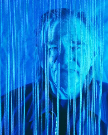 Portrait of an adult male with silver hair, posing behind blue vertical string lights, creating a textured, ethereal effect.