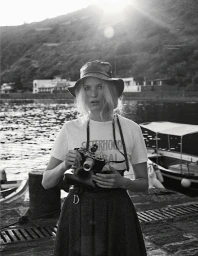 Black and white portrait of a woman wearing a bucket hat and t-shirt, holding a vintage camera by a harbor with boats.