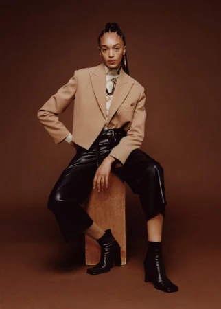Model with braided hair sitting on a wooden block wearing a camel blazer and black leather pants