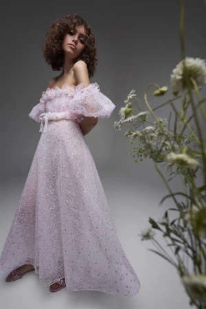 Model with curly hair in a pink off-the-shoulder gown standing against a grey backdrop with flowers in the foreground.