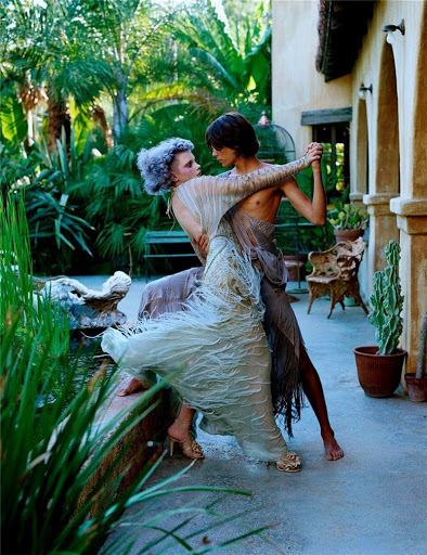 Two fashion models in flowing dresses dance barefoot outdoors in a lush tropical garden, featuring stucco architecture and terracotta pots.