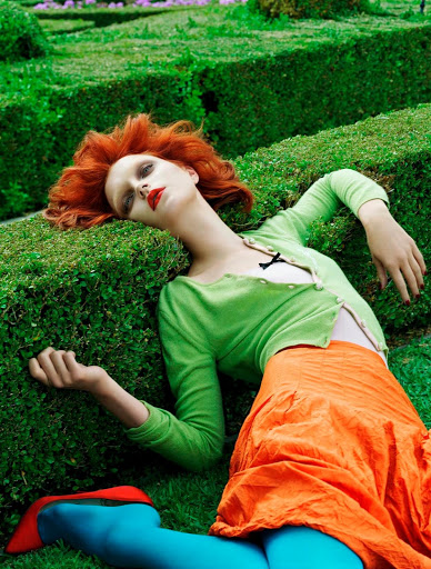 A model lies in a green hedge maze wearing an orange skirt, green sweater, blue tights, and red shoes in a fashion editorial.