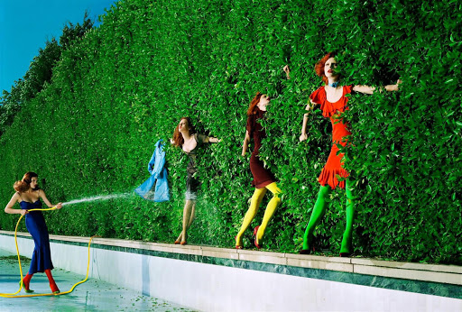 Fashion models in dresses pose behind a high green hedge, one holds a hose, the shot is a playful take on high fashion.