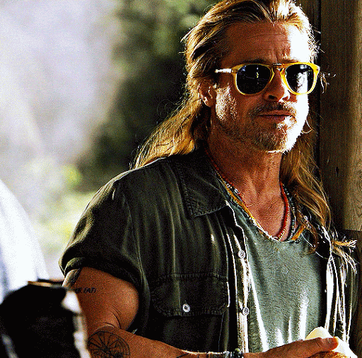 Close-up of Brad Pitt with long blond hair and sunglasses, wearing a green shirt and necklaces, standing outdoors during the daytime.