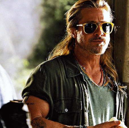 Close-up of Brad Pitt with long blond hair and sunglasses, wearing a green shirt and necklaces, standing outdoors during the daytime.