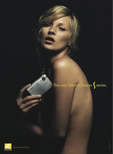 Kate Moss poses in a studio ad campaign for Nikon Coolpix, holding a silver compact camera. Dark background with yellow text.