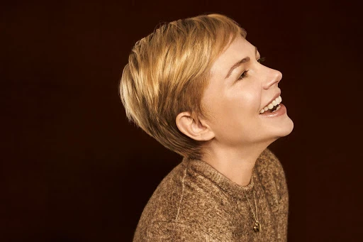 Profile portrait of smiling woman with short blonde hair wearing a brown knitted sweater against a warm dark background