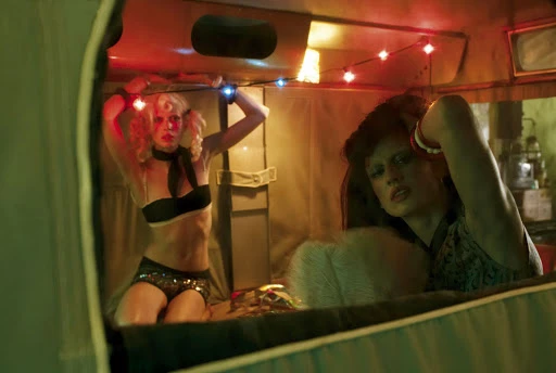 Two women in a vintage camper with retro outfits pose under string lights. One woman kneels with her arms raised. 