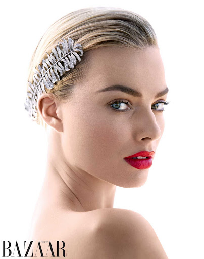 Close-up portrait of a blonde woman with red lipstick wearing a jeweled feather headpiece against a bright white backdrop in a beauty shot.