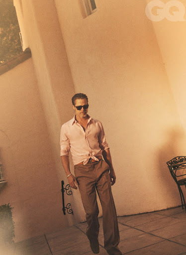 An adult male in sunglasses and pink collared shirt with brown pants stands outdoors on a tan surface with architectural details.