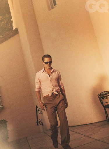 An adult male in sunglasses and pink collared shirt with brown pants stands outdoors on a tan surface with architectural details.