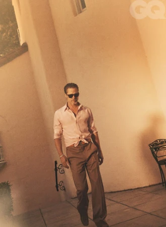 An adult male in sunglasses and pink collared shirt with brown pants stands outdoors on a tan surface with architectural details.