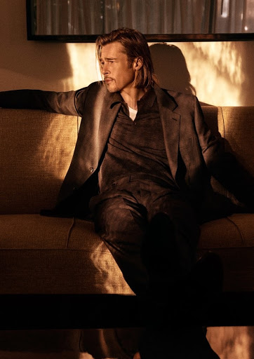Portrait of a blonde male celebrity wearing a suit, sitting on a sofa in a naturally lit, warm-toned indoor setting. 