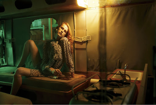 Red-haired adult woman in animal-print blouse poses seated in the interior of vehicle with sink, stove, yellow and green lighting.