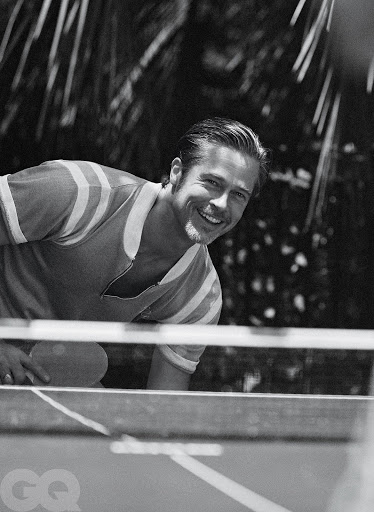 Black and white medium shot of a smiling adult male playing table tennis with a paddle, set against a blurred background of outdoor foliage.