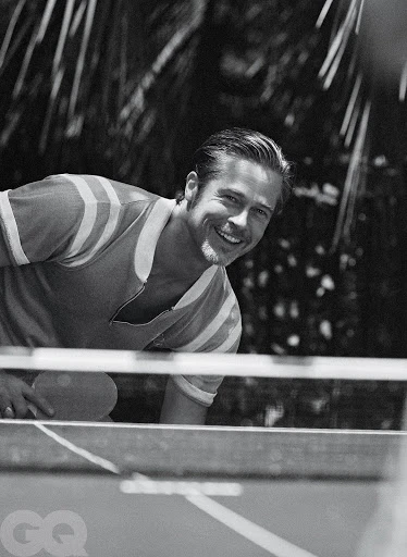Black and white medium shot of a smiling adult male playing table tennis with a paddle, set against a blurred background of outdoor foliage.