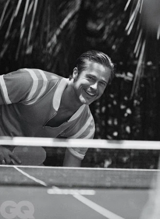 Black and white medium shot of a smiling adult male playing table tennis with a paddle, set against a blurred background of outdoor foliage.