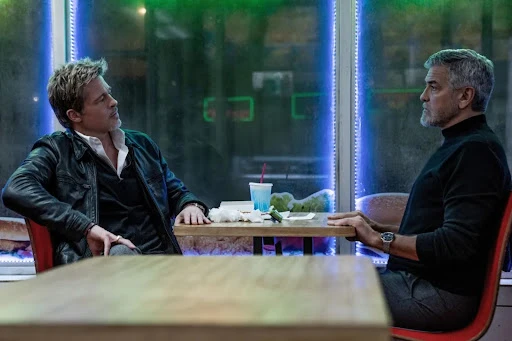 Two mature adult males sit at a table across from each other indoors. One man has blond hair and is wearing a black jacket.