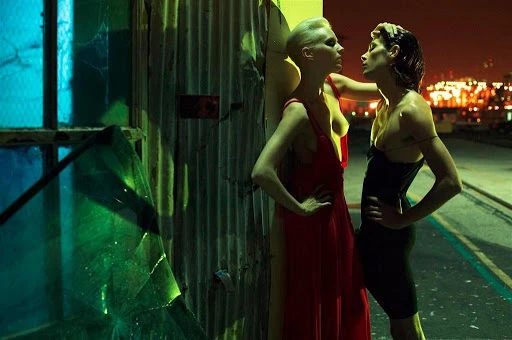 Two women posing outdoors at night against a corrugated wall, featuring dramatic neon lighting and cinematic grading by Johnathan Gale.