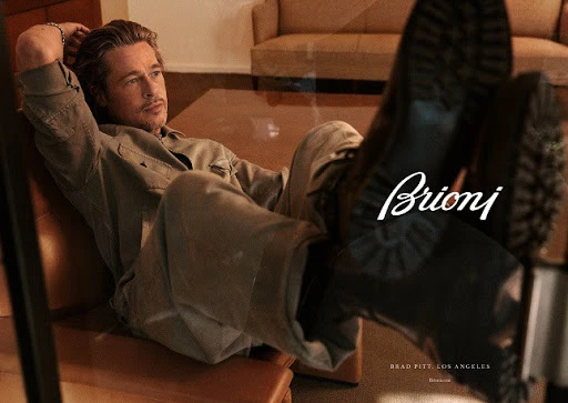 Brad Pitt lounges in an indoor setting, sporting a casual shirt and trousers, with a pair of black leather boots prominently displayed.