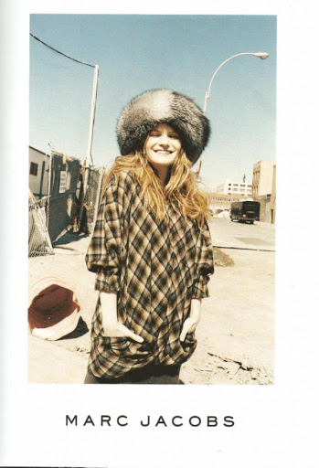 Outdoor shot of a woman smiling, wearing a plaid shirt and large fur hat, standing on a city street with buildings in the background.