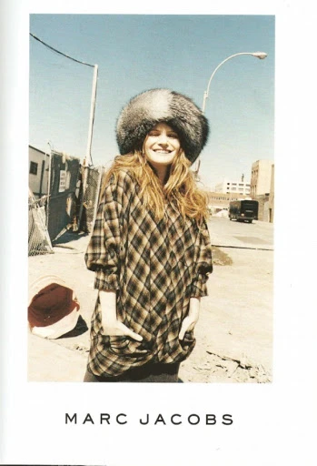 Outdoor shot of a woman smiling, wearing a plaid shirt and large fur hat, standing on a city street with buildings in the background.