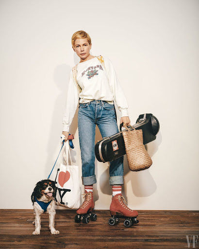 Michelle Williams on roller skates with a Cavalier King Charles Spaniel dog, carrying a tote bag, photographed against a white wall.