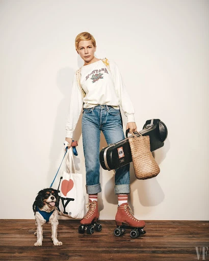 Michelle Williams on roller skates with a Cavalier King Charles Spaniel dog, carrying a tote bag, photographed against a white wall.