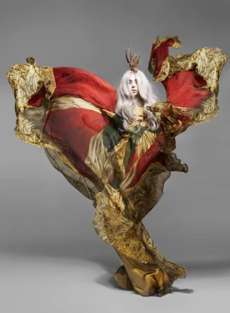 Woman in flowing red and gold patterned dress with crown poses against grey background in a studio photography fashion editorial.