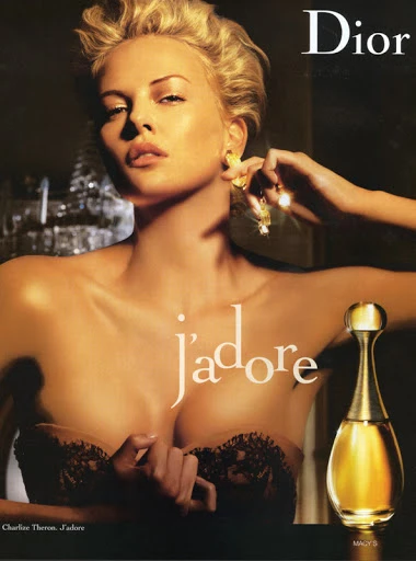 Charlize Theron for Dior perfume J'adore advertisement features a fair-skinned adult woman and a perfume bottle in a warm-toned, shallow focus.