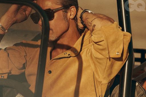 Adult male poses in a tan button-down shirt with sunglasses, showcasing wrist bracelets in a warm indoor setting with magazine logo.