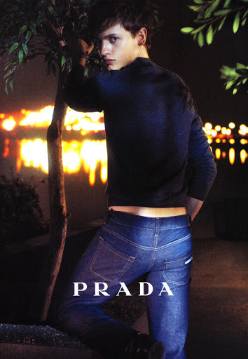 Male fashion model poses outdoors in a Prada denim ad, featuring dark blue jeans and top, next to a tree at nighttime with blurred lights.