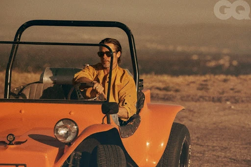 Brad Pitt driving an orange buggy in a desert landscape. He wears sunglasses and a yellow shirt in a vintage-style outdoor fashion shot.