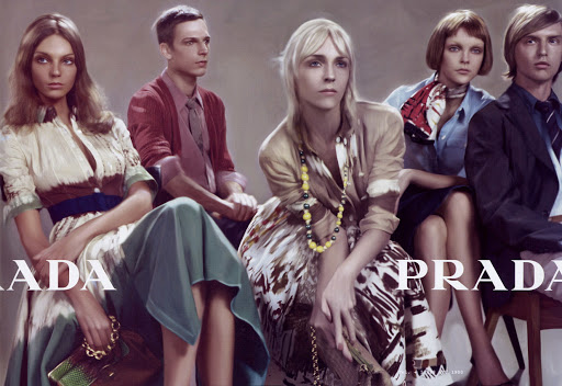 Fashion ad featuring five seated models—three women and two men—wearing outfits from a Prada collection, set against a neutral backdrop.