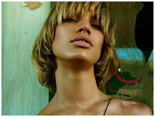 Close-up portrait of a fair-skinned adult female model with short blonde hair and light makeup posing against a textured studio backdrop.