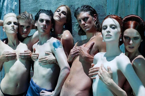 Group of seven models posing closely together against a textured wall with cool teal tones and dramatic lighting.