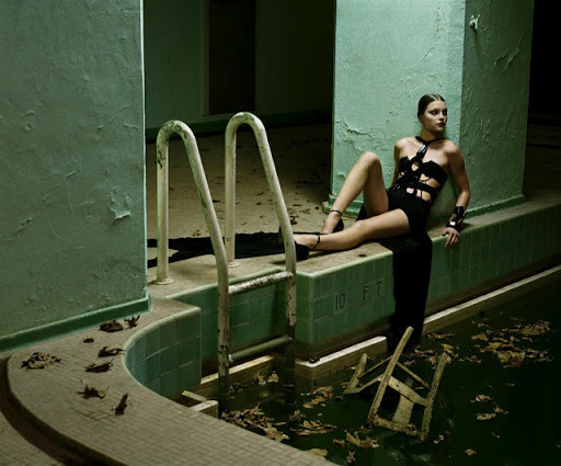 Model in black dress lounging by a murky abandoned indoor swimming pool with green tiled walls