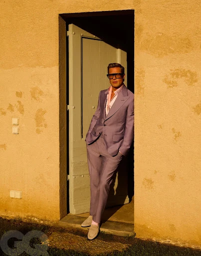 Mature man in a lavender suit and sunglasses leaning in the doorframe of a stone building, GQ logo at the bottom.