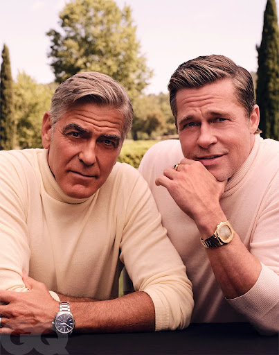 Two adult males, George Clooney and Brad Pitt, in a medium shot, both wearing watches and light colored sweaters in an outdoor setting.