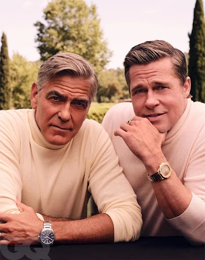 Two adult males, George Clooney and Brad Pitt, in a medium shot, both wearing watches and light colored sweaters in an outdoor setting.