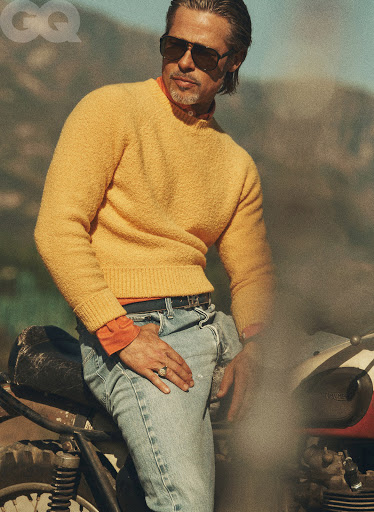 Portrait of male model, Brad Pitt, in sunglasses, orange shirt, and yellow sweater sitting on a vintage motorcycle in an outdoor setting.