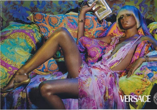 Versace ad features a reclining woman with blue hair, wearing a pink dress, gold sandals, and jewelry, holding a gold clutch, on patterned cushions.