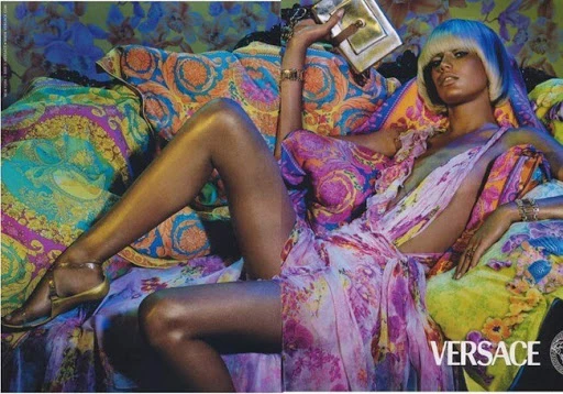 Versace ad features a reclining woman with blue hair, wearing a pink dress, gold sandals, and jewelry, holding a gold clutch, on patterned cushions.