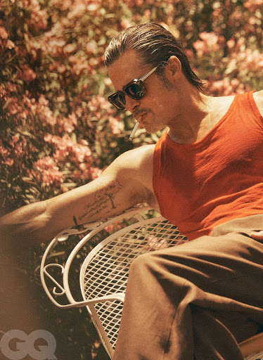 Adult male with wet slicked hair, wearing an orange tank top and sunglasses, sitting on a white chair, outside amongst plants and trees.