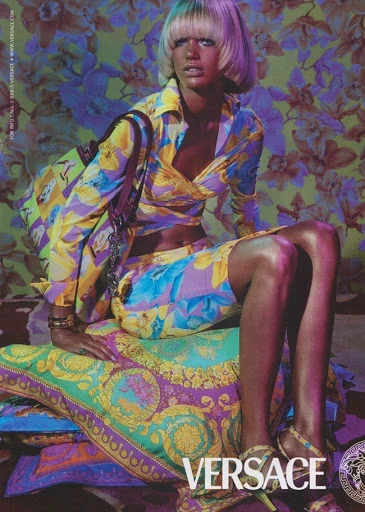 A stylish woman with a blonde bowl cut sits on patterned pillows in a matching floral Versace outfit, posing against a floral backdrop.