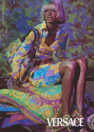 A stylish woman with a blonde bowl cut sits on patterned pillows in a matching floral Versace outfit, posing against a floral backdrop.