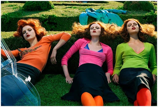 Three red-haired women in colorful sweaters and dark skirts lying on grass in garden setting with formal topiary and clear plastic chair.