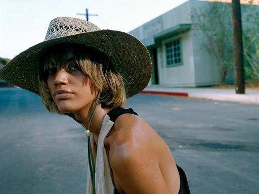 Woman wearing a wide-brimmed straw hat and a halter top on an empty street with a light-colored building in the background.