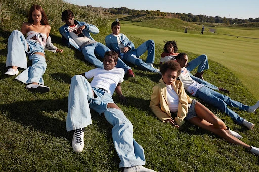 Group of seven young diverse adults in denim clothing casually lounging on a grassy hill with a golf course visible in the background on a sunny day.